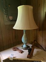 Photo of vintage table lamp with painted light blue base and cream fabric lampshade on a white surface against wood panel wall