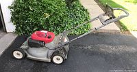 Side view of Honda walk-behind lawn mower showing red engine cover and gray deck with four wheels, handle with rust spots visible.