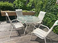 Full patio set of round table and 4 chairs displayed outside on a wooden deck with plants in background under sunlight.