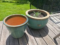 Two teal ceramic planters on wooden deck, one empty and one with soil and plants.