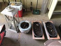 Overview photo of the set including two rustic clay planters with plastic inserts and soil, one floral ceramic planter with soil, and a tall gray metal candle lantern.