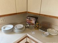 Photo showing white china plates stacked along with decorative bowls, a ceramic decanter with floral design, a muffin tin and loaf pan cake tin, a small metal bowl, glass jars with lids, and a new-in-box wooden salad set.
