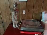 Glass serving plates in original boxes with large pottery statue on a wooden surface against paneled wall.