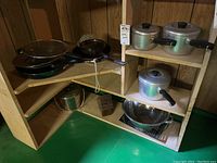 Corner shelf with various aluminum pots and pans including Lagostina brand, metal strainer, and wooden cutting block below.