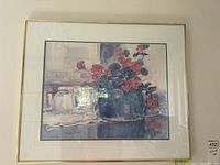 Framed floral print with red flowers in a blue vase, on a surface with other ceramic vessels, metal frame with white mat.