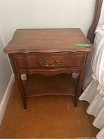 Front angle view of one wooden side table showing drawer with metal pull, scalloped edges, and lower curved shelf.