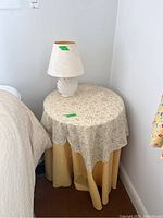 Round table covered with beige floral fabric skirt and vintage lamp with cream lampshade on top, placed next to bed.
