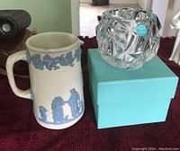 Tiffany & Co crystal candle holder displayed on its Tiffany blue box next to the Wedgwood pitcher on a red surface.