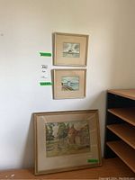 Three watercolour art pieces framed: two smaller square coastal scenes stacked on the wall, one larger rectangular landscape on the floor leaning against wall