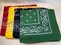Four stacks of bandannas in different colors, folded and displayed on a white surface. Colors from front to back: green, navy blue, yellow, red.