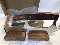 Photo shows two new chef cutting utensils with curved stainless steel blades and dark wooden handles, accompanied by two dark wooden rests. Items are in a cardboard box with bubble wrap inside.