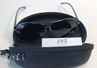 J+S men’s aviator sunglasses resting on a black zippered hard case with J+S logo and an informational card inside case