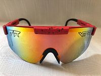 Front view of men's sunglasses showing large gradient mirrored lens and red adjustable arms with black speckles.