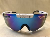 Close up photo of men's sports sunglasses with a blue mirrored visor style lens and white frame arms that have black speckled pattern. The arms have visible adjustment mechanisms. The lens shows Viper logos in blue and red on each side.