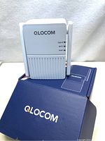 White QLOCOM WiFi extender device with single antenna displayed on top of the branded blue box, showing front panel with signal, WPS, and power indicators.