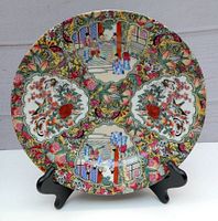 Front view of the vintage Chinese Famille Rose porcelain plate displayed on a black stand showing detailed hand-painted Mandarin figures, floral and architectural designs.