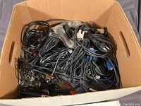 Photo of a cardboard box filled with a tangled assortment of black cables including Cat 6 patch cords with blue connectors, USB cords, and power supply cables.