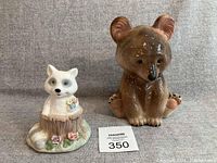 Photo showing the full lot with the ceramic raccoon music box on a tree stump, decorated with flowers, next to a brown ceramic bear cub figurine, both sitting on a neutral backdrop.