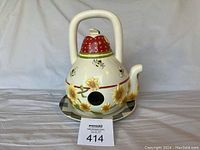 Front view of the ceramic teapot bird house showing the spout, handle, red lid with white polka dots and small ceramic detail on top, bees and yellow flower decoration, with the attached plate underneath.