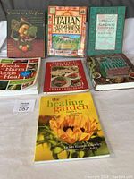 Seven cooking and healing books laid on a surface, titles visible including Cooking for a New Earth, Italian Farmhouse Cookbook, Midwest Gardener's Cookbook, and The Healing Garden.