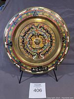 Front view of hand painted brass plate displaying detailed colorful Aztec calendar design, prominently showing the inscription 'MEXICO'. Plate is round, 11 inches in diameter, with vibrant colors.