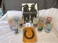 Group photo showing the book, wooden horseshoe wall hanging, and 10 Kentucky Derby glasses arranged on fabric background.