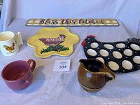 Photo of vintage kitchen group including 'Bless this kitchen' sign, yellow chicken platter, black hen egg server, Oneida creamer, white chicken mug, and red cup