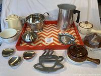 Photo shows assorted kitchen items arranged on a red and cream chevron patterned serving tray, including teapots, a colander, metal dishes, and a brown handled pot.