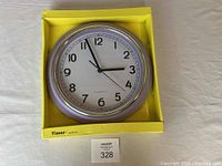 Full view of the wall clock in its yellow packaging showing the round shape and silver rim.