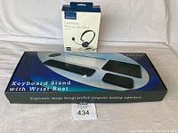 Photo shows the keyboard stand with wrist rest packaging alongside the boxed landline phone headset on a white cloth background.