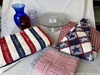 Photo of three folded patriotic tablecloths, placemats and glass vases with cake plate on white cloth background.