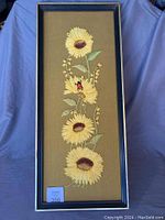 Full front view of the framed vintage yarn embroidered wall art showing four sunflowers vertically aligned on brown fabric background framed in dark blue wood.