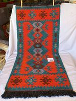 Full view of woven textile with geometric and diamond patterns in red, blue, and brown, with black fringe at ends.