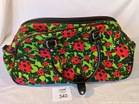 Front view of the zippered travel bag showing ladybug print, two black handles with silver hardware, and a snap closure tab at base of handles.