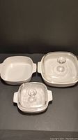 Top-down view of three CorningWare square baking dishes. Two have clear glass lids, medium size dish missing lid.
