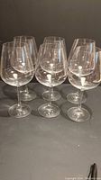 Set of six clear crystal wine glasses with long stems and wide bowls, arranged in two rows of three on a dark surface.