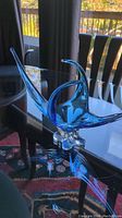 Full view of blue and clear glass art sculpture on reflective black surface with outdoor background.