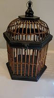 Hexagonal bird cage closed, showing bamboo slats and black wooden frame with top finial and hanging ring.