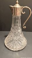 Full view of the vintage cut glass decanter showing the clear intricate diamond and starburst cut glass bottom and tapered neck with silver plated top and curved spout handle.