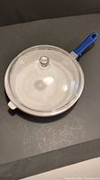 Top view of the white skillet with a clear glass lid and a blue removable handle.