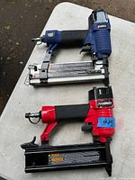 Photo of two pneumatic air nailers placed side by side on a white surface. Top nailer is Campbell Hausfeld in blue and black, bottom nailer is Husky in red and black with blue tape label.