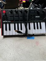 Front view of AKAI MPK Mini Play keyboard showing 25 keys, buttons, knobs, red joystick, USB cable attached on a table.