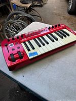 Red Behringer MIDI keyboard with 25 keys shown from angled top-left view, placed on a grey table with wheels visible in background.