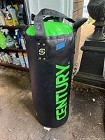 Photo showing a black and green Century punching bag standing upright on a concrete floor with cleaning supplies in the background.
