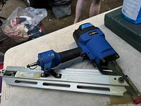 Left side angled view of blue and black Campbell Hausfeld air framing nailer on white table outdoors, showing metal nail magazine with protective tape on the nose.