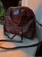 Front angled view showing brown leather purse with gold-tone studs and shoulder strap placed on a marble surface near a window.