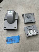 Photo shows three Nintendo 64 accessory items on a gray surface: a Rumble Pak, a Controller Pak, and a Memory Card. A small blue tag with handwritten markings is also visible.