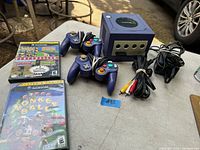 Nintendo GameCube console, two controllers, two game cases, and cables laid on a table outdoors.