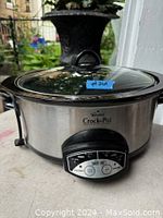 Stainless steel Rival Crock-Pot slow cooker with digital control panel and glass lid shown outside on a surface.
