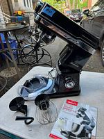 Photo of Sunbeam Mixmaster Pro stand mixer with attachments displayed around it on a table, showing the black mixer body, chrome accents, and attachments including dough hook, beater blade, and wire whisk.